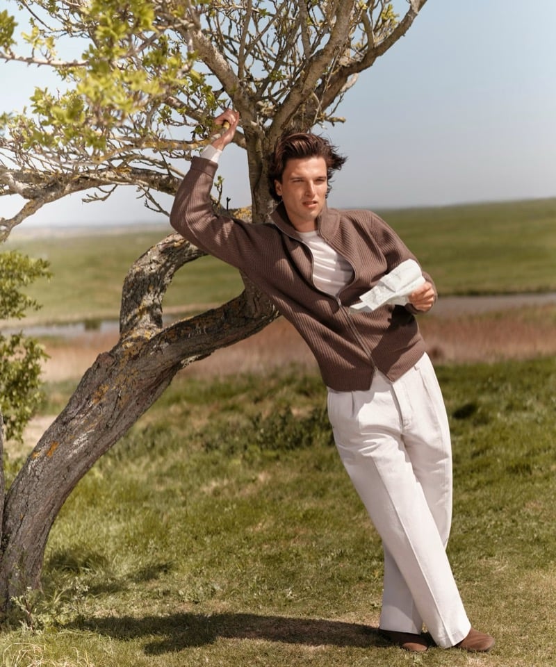 Kit Clarke leans against a bare tree in open countryside, wearing a brown ribbed zip-through knit over a striped T-shirt with pale wide-leg trousers and suede loafers for John Smedley