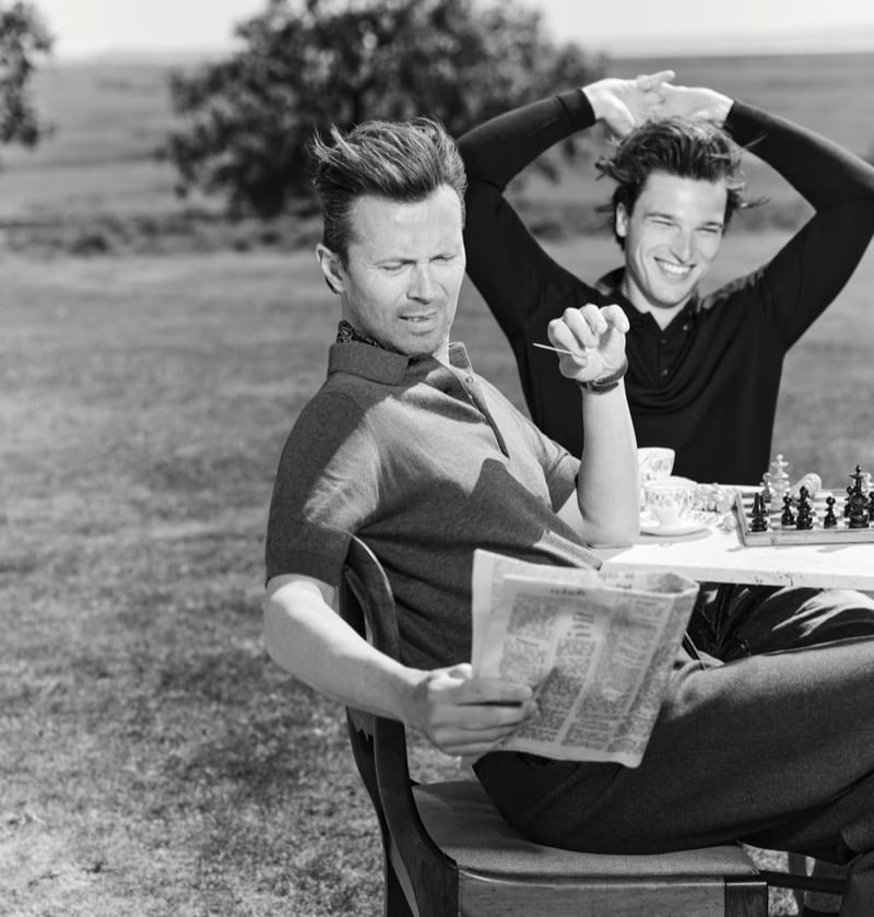 Black and white image of Robert Montgomery seated with a folded newspaper in a piqué polo shirt while Kit Clarke stretches his arms overhead behind him, a chess board and teacups on the table between them in a field
