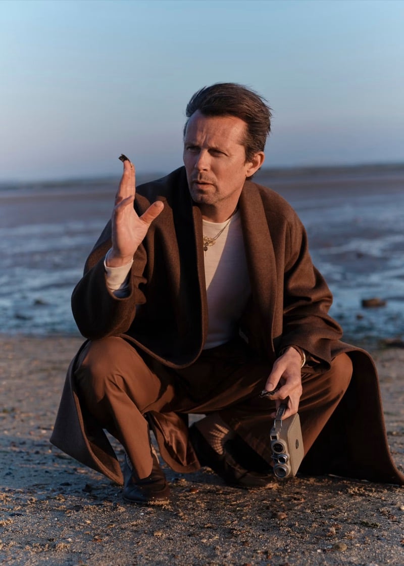 Robert Montgomery crouches on a beach at dusk in a brown shawl-collar wrap coat over a light T-shirt with a gold pendant necklace, holding a vintage cine camera