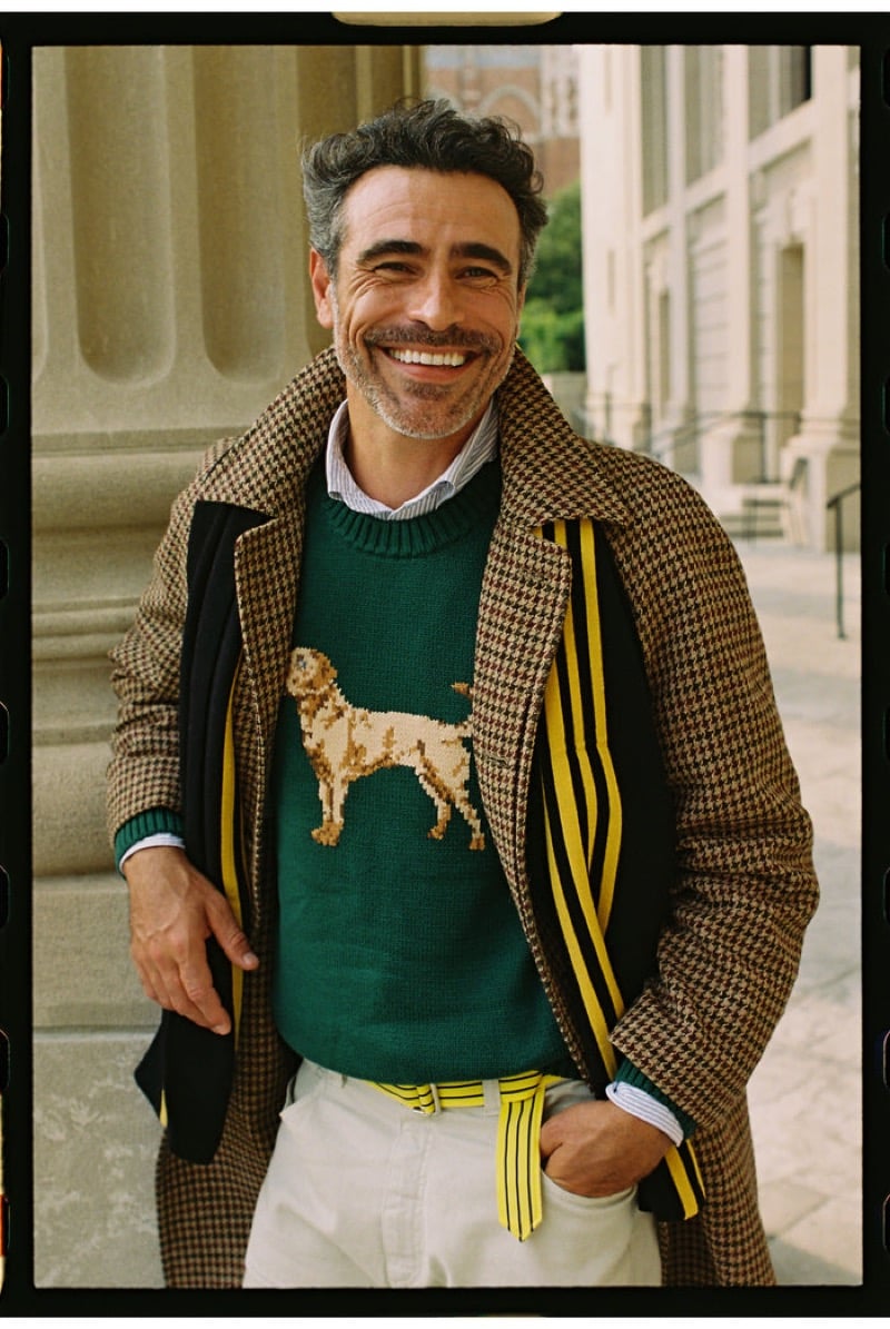 A smiling model wears a brown houndstooth overcoat over a green sweater knit with a dog motif, a striped shirt, and a striped scarf with off-white trousers, for J. Press fall-winter 2026