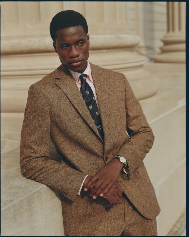 A model leans against a stone column base in a brown Donegal tweed suit worn with a striped shirt and a crest-motif tie, for J. Press fall-winter 2026