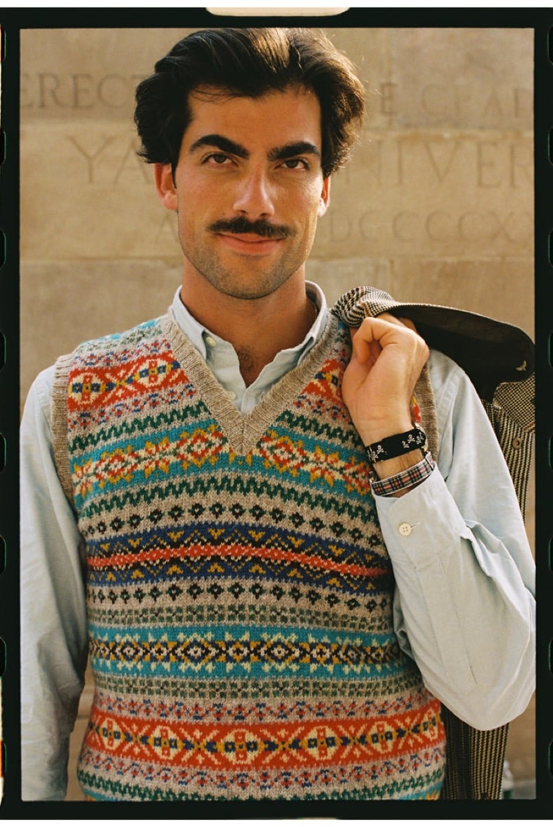 A model holds a houndstooth jacket over one shoulder, wearing a multicolor Fair Isle sweater vest over a light blue Oxford shirt in front of carved Yale University stone, for J. Press fall-winter 2026