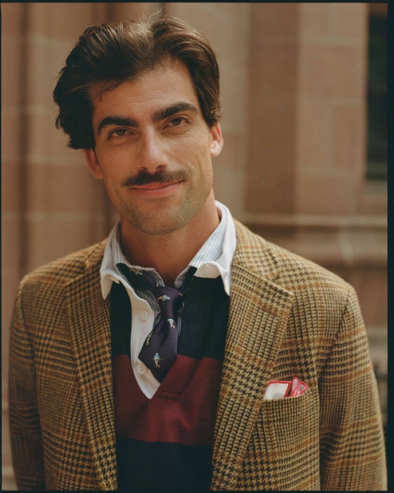 A close portrait of a model in a tan glen-check tweed jacket over a burgundy color-block rugby polo with a patterned tie and a paisley pocket square, for J. Press fall-winter 2026