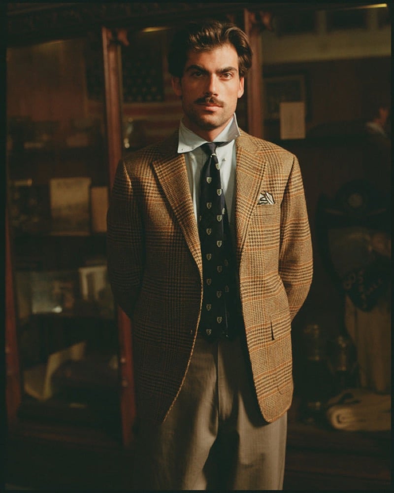 A model wears a glen-check tweed sport coat with a light blue Oxford shirt, a crest-motif tie, and gray trousers in a wood-paneled trophy room, for J. Press fall-winter 2026