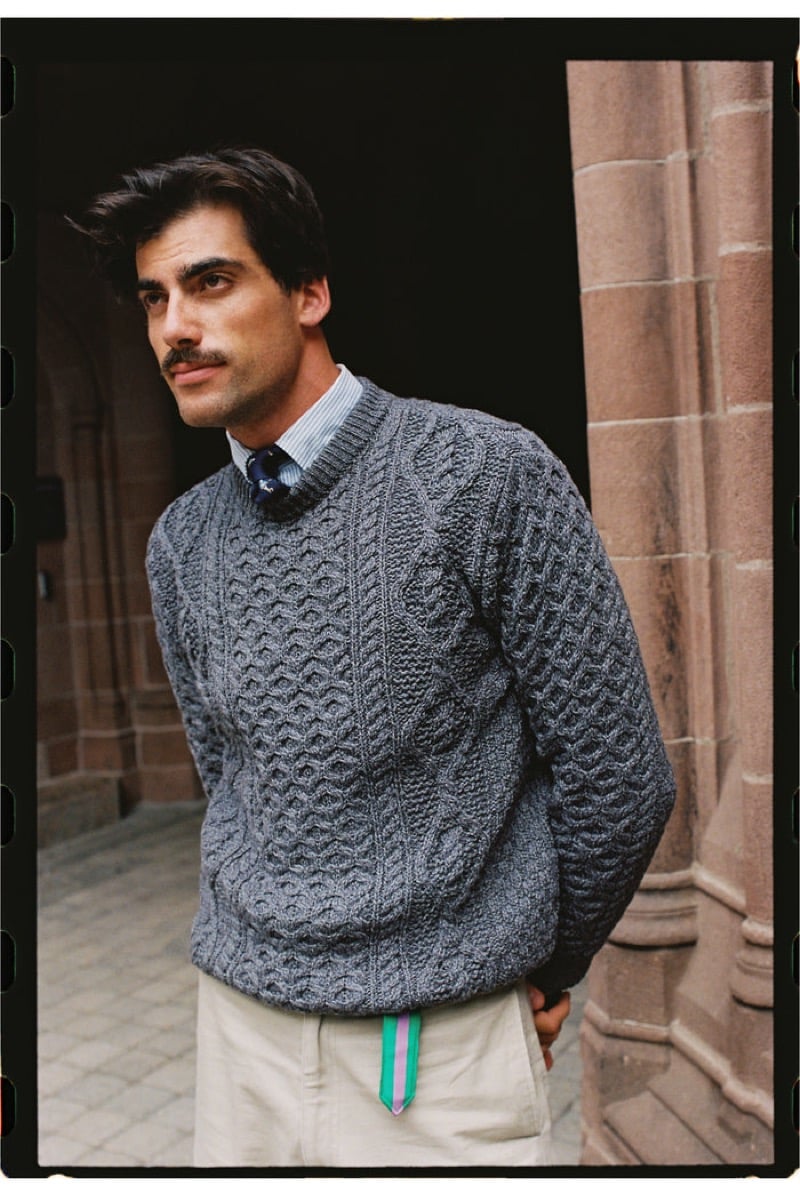 A model wears a gray Aran cable-knit sweater over a striped shirt with a patterned tie and stone trousers with a pink and green striped belt, in a stone archway, for J. Press fall-winter 2026