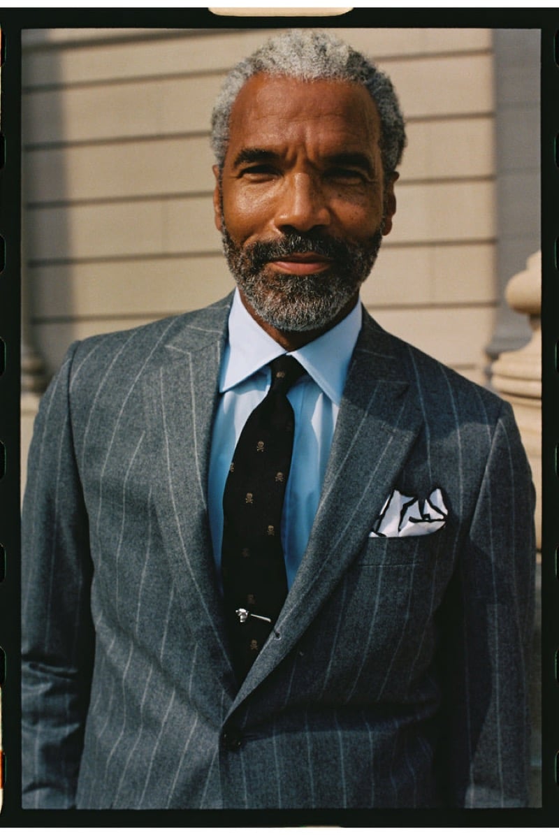 An older model with a gray beard wears a gray chalk-stripe flannel suit with a light blue shirt and a skull-motif tie with a tie pin, outdoors by a column, for J. Press fall-winter 2026