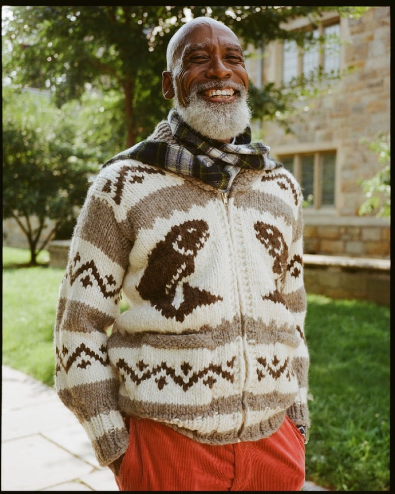 A smiling older model wears a hand-knit wool zip sweater with animal motifs and geometric bands over a plaid flannel shirt with rust corduroy trousers, outdoors on campus, for J. Press fall-winter 2026