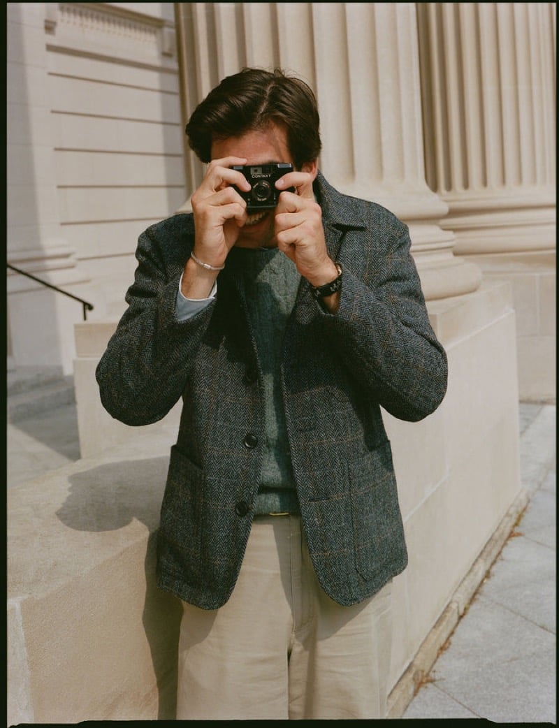 A model raises a film camera to his face, wearing a herringbone tweed jacket with a windowpane check over a green sweater with stone trousers, on stone steps, for J. Press fall-winter 2026