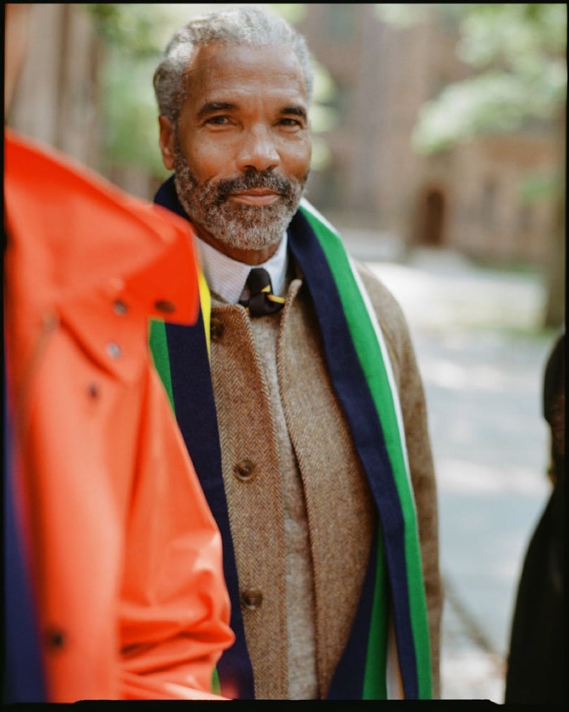 An older model with a gray beard wears a herringbone tweed suit with a dark bow tie and a green and blue college scarf, standing beside a figure in a bright orange rain jacket, for J. Press fall-winter 2026