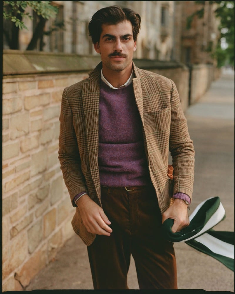 A model wears a tan houndstooth tweed jacket over a heathered purple sweater with brown corduroy trousers, holding a striped scarf on a campus sidewalk, for J. Press fall-winter 2026