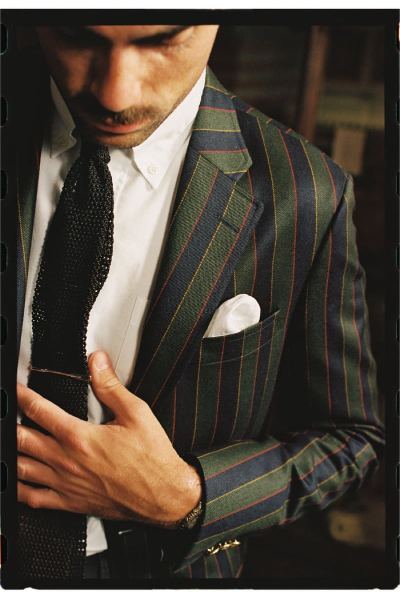 A close view of a model adjusting a knit tie, wearing a boldly striped wool jacket in green and blue with red and yellow stripes over a white shirt, for J. Press fall-winter 2026