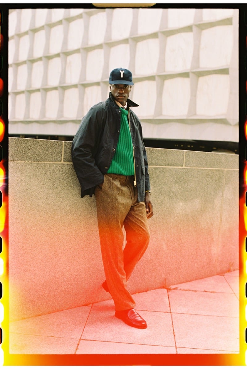 A model leans against a wall in a dark waxed cotton jacket over a green ribbed sweater with brown flecked wool trousers, a wool letter cap, and leather loafers, for J. Press fall-winter 2026