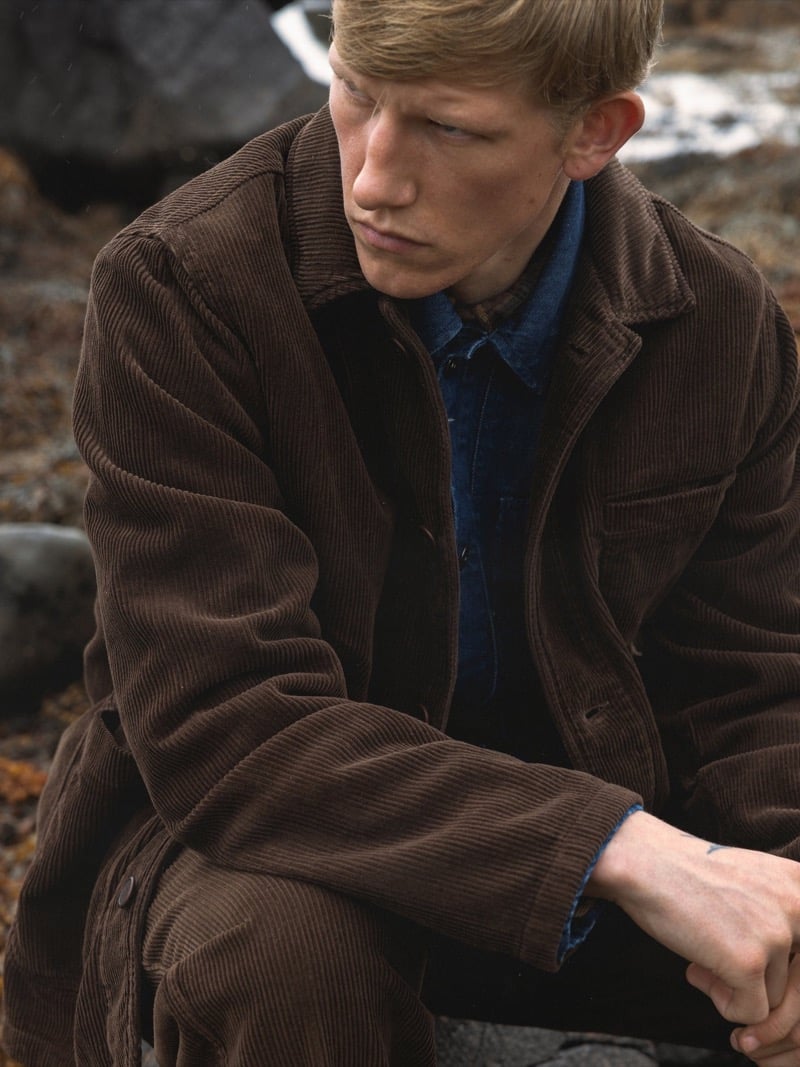 Connor Newall crouches among coastal rocks with patches of snow behind him, wearing a brown corduroy chore jacket over an indigo denim shirt