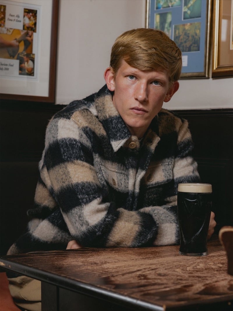 Connor Newall sits at a pub table with a pint of stout in front of him, wearing a brushed wool overshirt in a large cream and charcoal buffalo check, framed photographs on the wall behind him