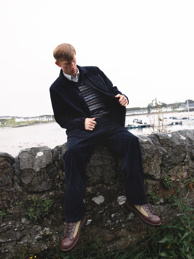 Connor Newall sits on a stone harbor wall opening a corduroy chore jacket over a Fair Isle sweater vest, with matching corduroy trousers and brown boots