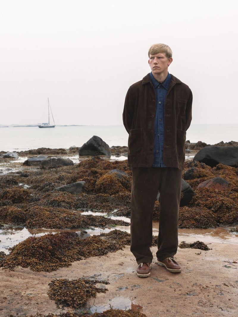 Connor Newall stands on a seaweed-covered beach in a brown corduroy overshirt over an indigo denim shirt, with brown corduroy trousers and a sailboat on the water behind him