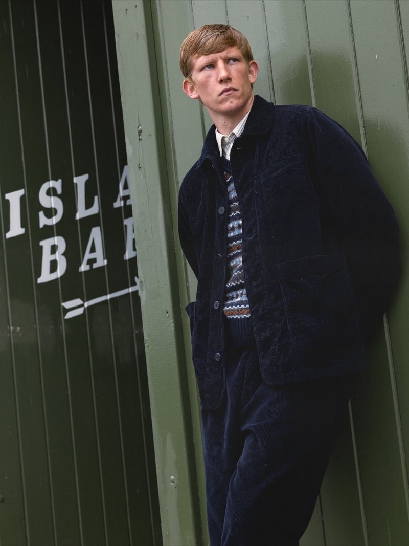 Connor Newall leans against a green wooden wall lettered Islay Bar in a corduroy chore jacket and matching wide trousers over a Fair Isle sweater vest and a striped shirt