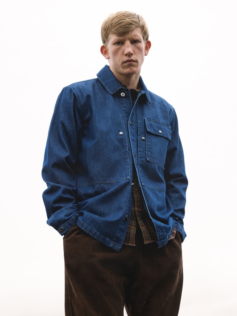 Connor Newall stands against a white studio backdrop in an indigo denim chore jacket over a brown plaid flannel shirt, with brown corduroy trousers and his hands in his pockets