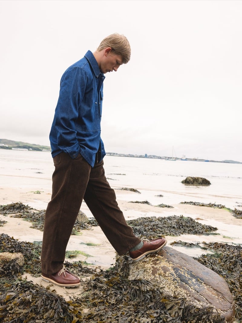 Connor Newall walks across seaweed-covered rocks on a beach in profile, wearing an indigo denim overshirt with brown corduroy trousers and brown moccasin boots