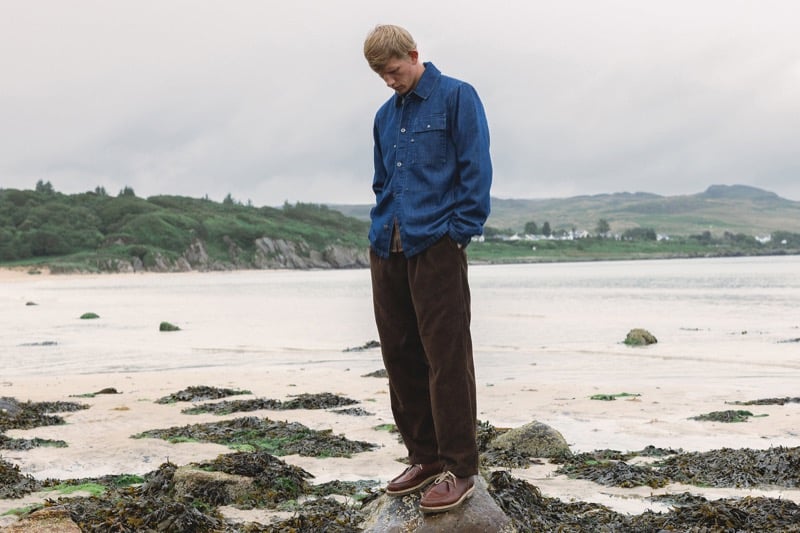 Connor Newall stands on a rock on the beach looking down, wearing an indigo denim shirt with brown corduroy trousers and brown boat shoes