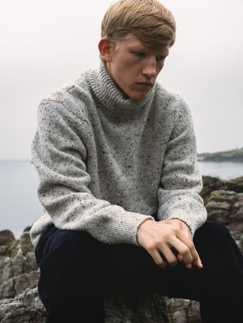 Connor Newall sits on rocks by the sea in a flecked Donegal wool rollneck sweater with dark corduroy trousers