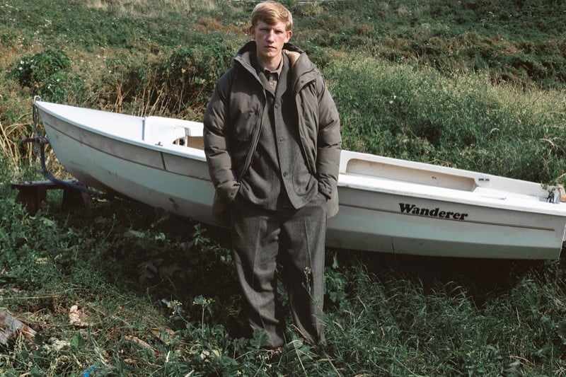 Connor Newall stands in a grassy field in front of an overturned white dinghy marked Wanderer, wearing a padded down jacket over a tailored jacket with pleated trousers, his gloved hands in his pockets
