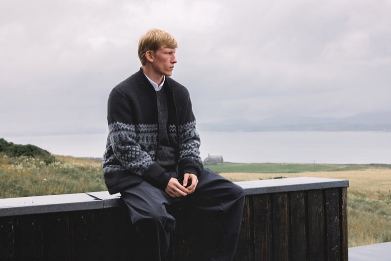 Connor Newall sits on a dark wood ledge overlooking the sea in a black and gray Fair Isle wool bomber over a dark sweater and a white shirt, with wide dark trousers