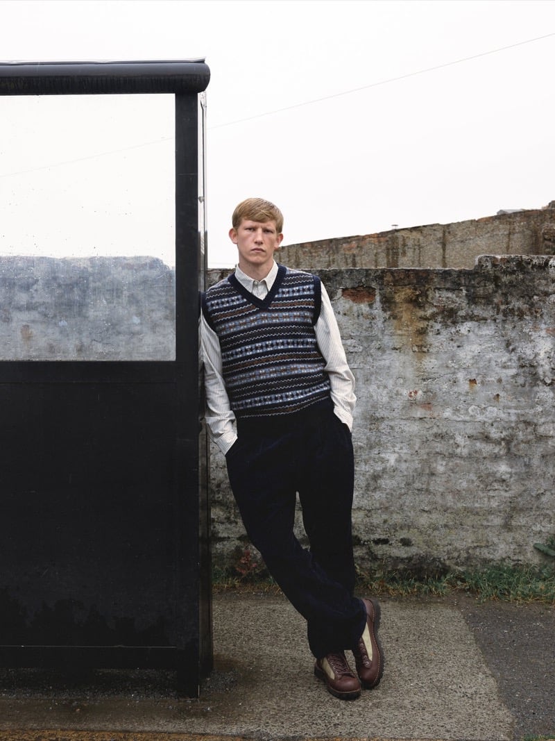 Connor Newall leans against a glass bus shelter in a patterned Fair Isle sweater vest over a striped button-up shirt, with dark wide-wale corduroy trousers and brown lace-up boots