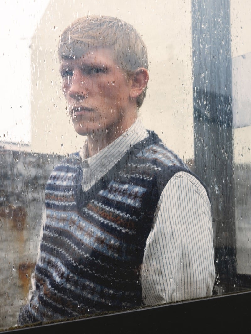 Connor Newall seen through a rain-streaked window wearing a Fair Isle sweater vest over a striped shirt