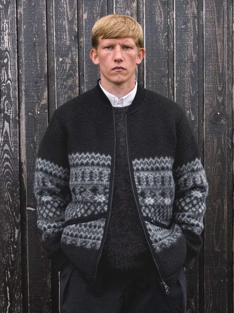 Connor Newall stands against a charred black timber wall in a wool bomber jacket with a Fair Isle pattern band, worn over a charcoal sweater and a white shirt