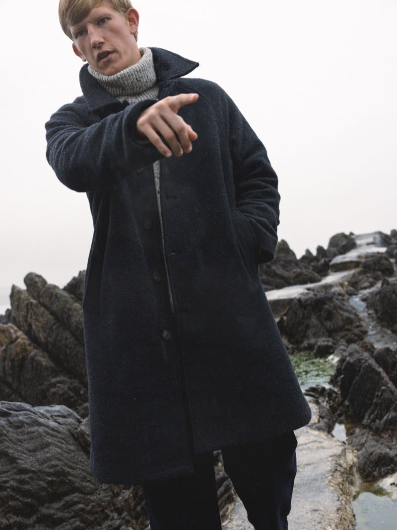 Connor Newall points while standing among coastal tide pools, wearing a dark flecked wool car coat with a stand collar over a gray rollneck sweater