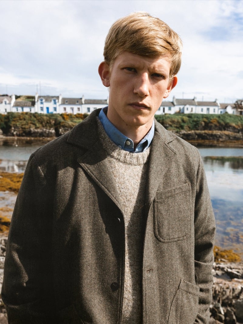 Close portrait of Connor Newall standing by the water with white cottages behind him, wearing a herringbone tweed workwear jacket over a flecked gray crewneck sweater and a blue chambray shirt