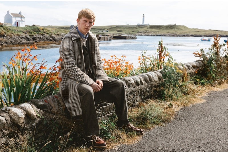 Connor Newall sits on a stone harbor wall with a lighthouse behind him, wearing a houndstooth check overcoat over a dark jacket with gray trousers and brown boat shoes