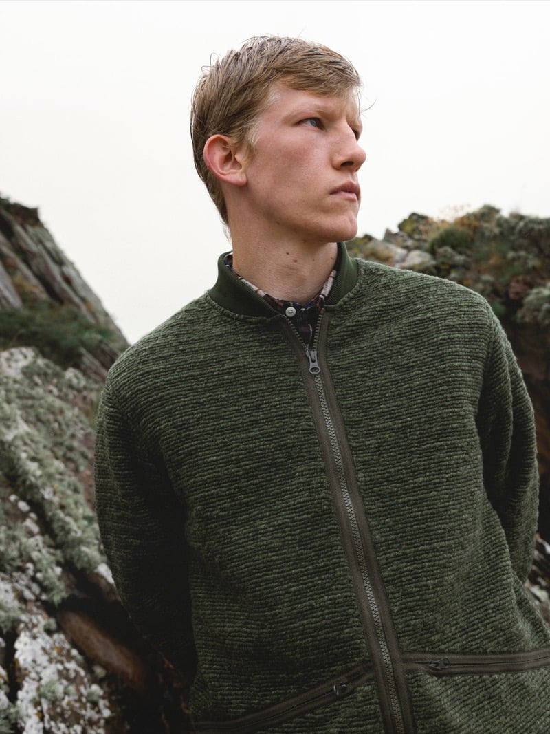 Connor Newall looks off to the side against lichen-covered cliffs, wearing an olive marled full-zip knit jacket over a check flannel shirt