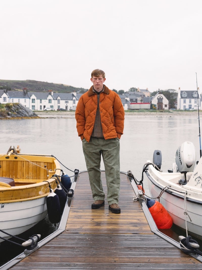 Connor Newall stands on a wooden jetty between moored boats in a burnt-orange quilted down jacket over a brown knit, with olive trousers and a village behind him