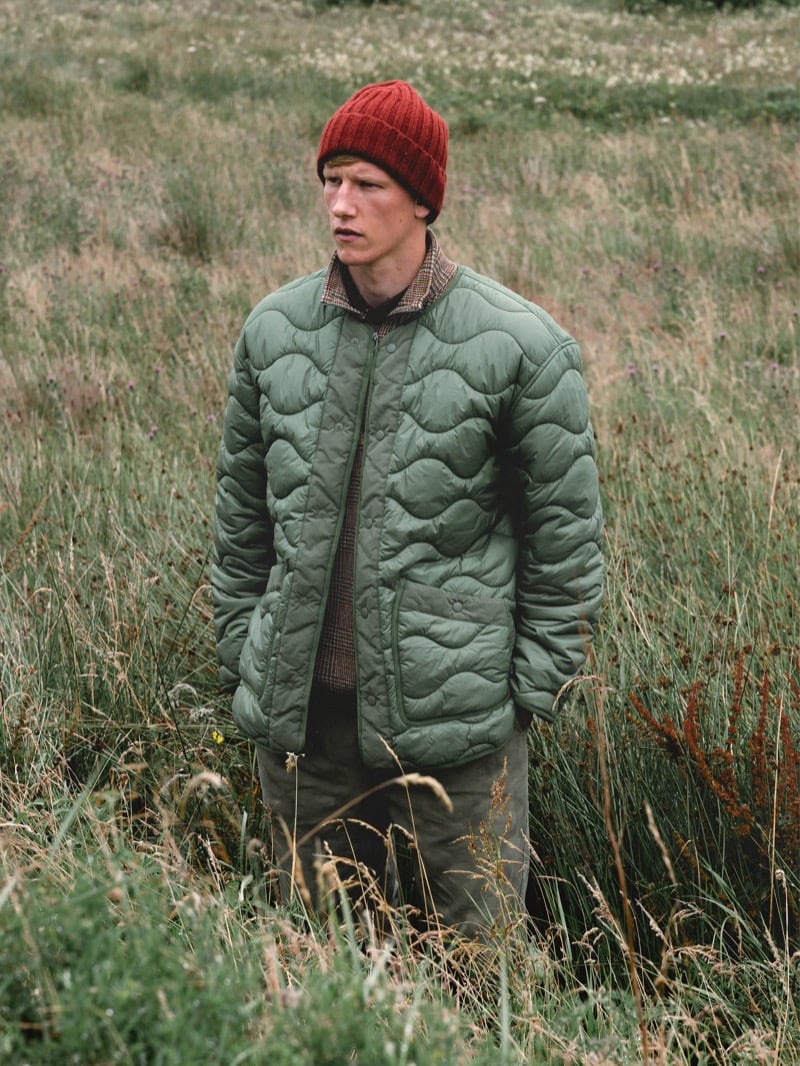 Connor Newall stands in a field of tall grass wearing a rust-colored ribbed beanie and a sage green onion-quilted liner jacket over a check overshirt and a brown knit, with olive trousers