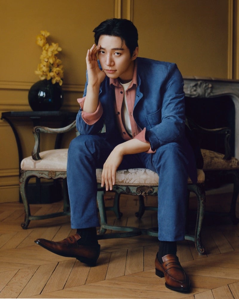 Lee Jun-ho seated on an antique upholstered bench in a blue corduroy suit worn over a pink shirt and white t-shirt, with brown leather loafers and dark socks, ochre walls and yellow orchids behind him