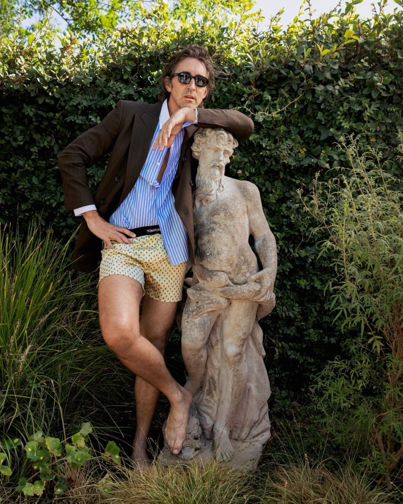 Lee Pace leaning on a garden statue in sunglasses, a brown Tom Ford jacket, striped shirt, and printed boxer shorts