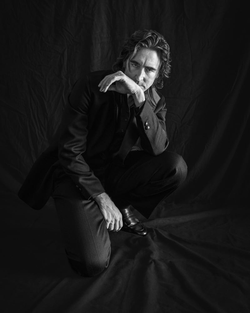 Black-and-white portrait of Lee Pace kneeling in a Zegna suit and loafers, hand resting at his chin