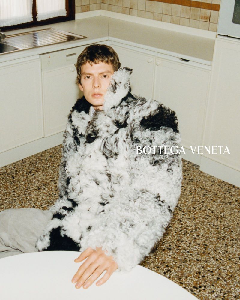 Leon Dame seated on a terrazzo kitchen floor in a curly shearling coat, one hand resting on a round table