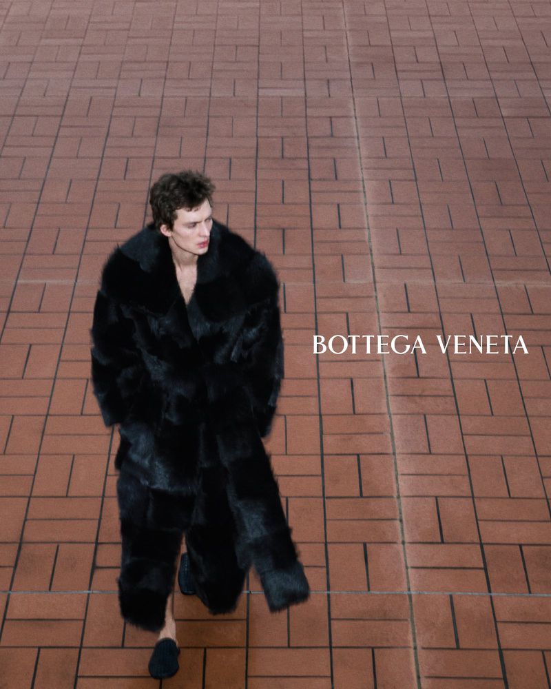 Leon Dame walks across a brick plaza in a floor-length shearling coat and woven slip-on shoes