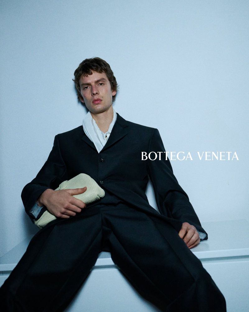 Leon Dame leans back against a wall in a long tailored coat and wide trousers, holding a woven leather clutch