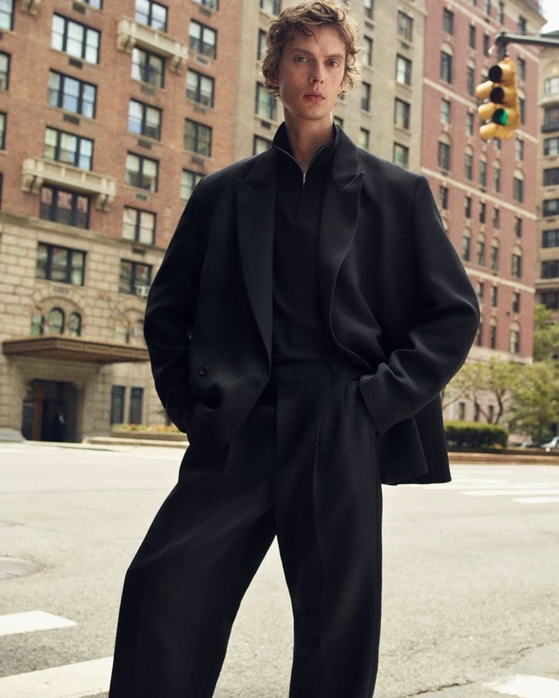 Leon Dame stands at a crosswalk with his hands in his pockets in an oversized blazer, a zip-collar knit, and wide pleated trousers, for COS's fall 2026 campaign