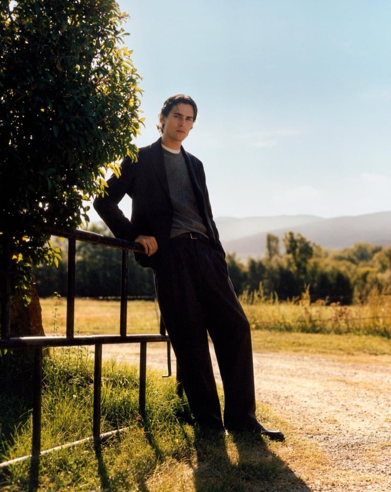 Liam Kelly in a blazer, gray sweater, and pleated trousers leaning on a metal gate in the countryside