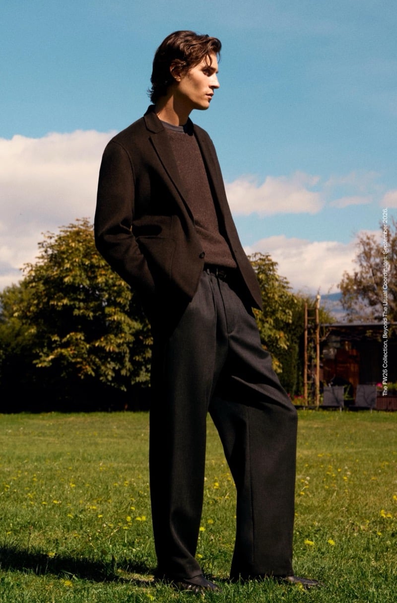 Liam Kelly in profile wearing a soft blazer, crewneck sweater, and wide-leg trousers on a sunny lawn