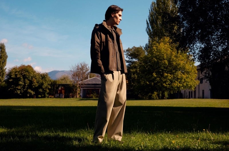 Liam Kelly in a field jacket and light pleated trousers standing on a sunlit lawn