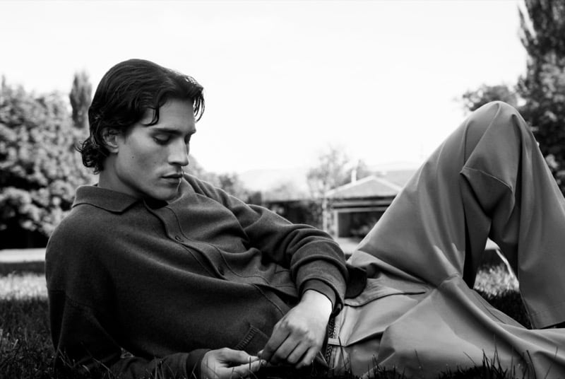 Black-and-white photo of Liam Kelly lying on the grass in a knit polo and wide trousers