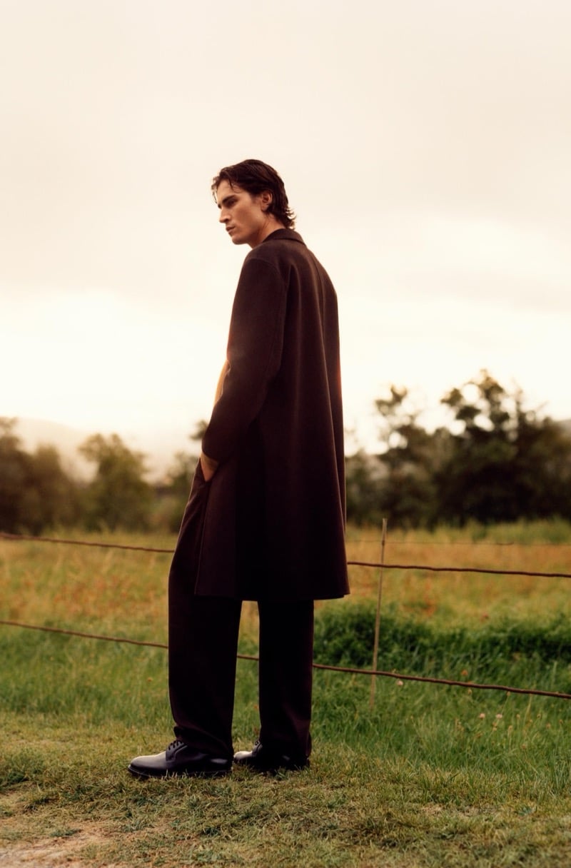 Liam Kelly in a long coat, trousers, and leather shoes standing by a wire fence in a field