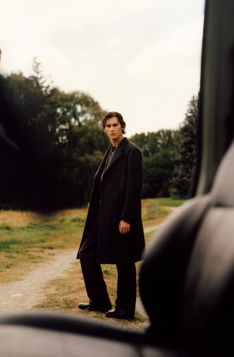 Liam Kelly in a long overcoat on a gravel road, seen through an open car door