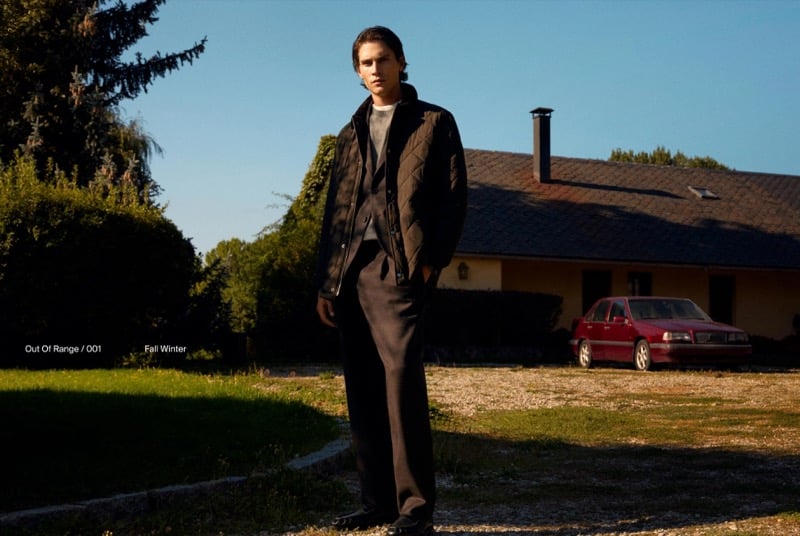 Liam Kelly in a quilted jacket and pleated trousers on a lawn in front of a country house and a red car